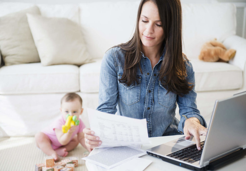 Mother is working with laptop while daughter (6-11 months) is sitting in background